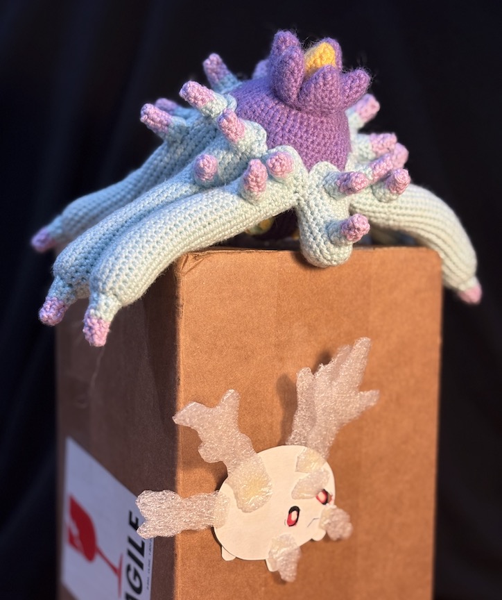 Crochet mareanie laying on a box, tentacles draped over the sides, as she looks down on a mostly-2D Galarian corsola made of cardstock and styrofoam