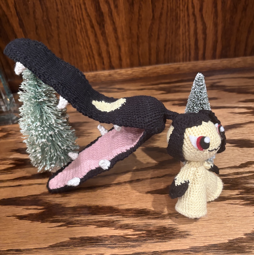 crocheted mawile in front of tiny evergreen trees with her chomp ponytail open