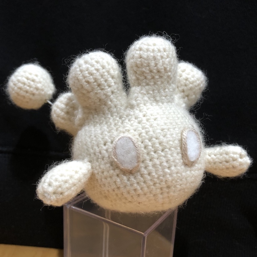 crocheted milcery at an angle and perched on a clear plastic box. She's stylized to have no mouth