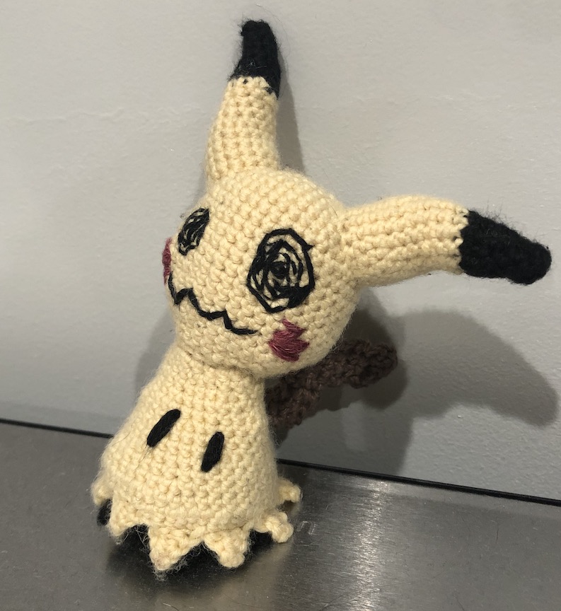 The pale crocheted mimikyu looking shyly at you, revealing more of the scribbled pikachu face on its disguise