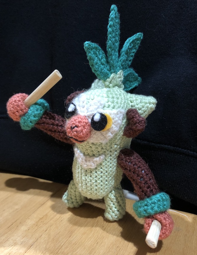 Crocheted thwackey from an angle, one arm raised with his stick, ready to drum, the other down, just having struck the ground
