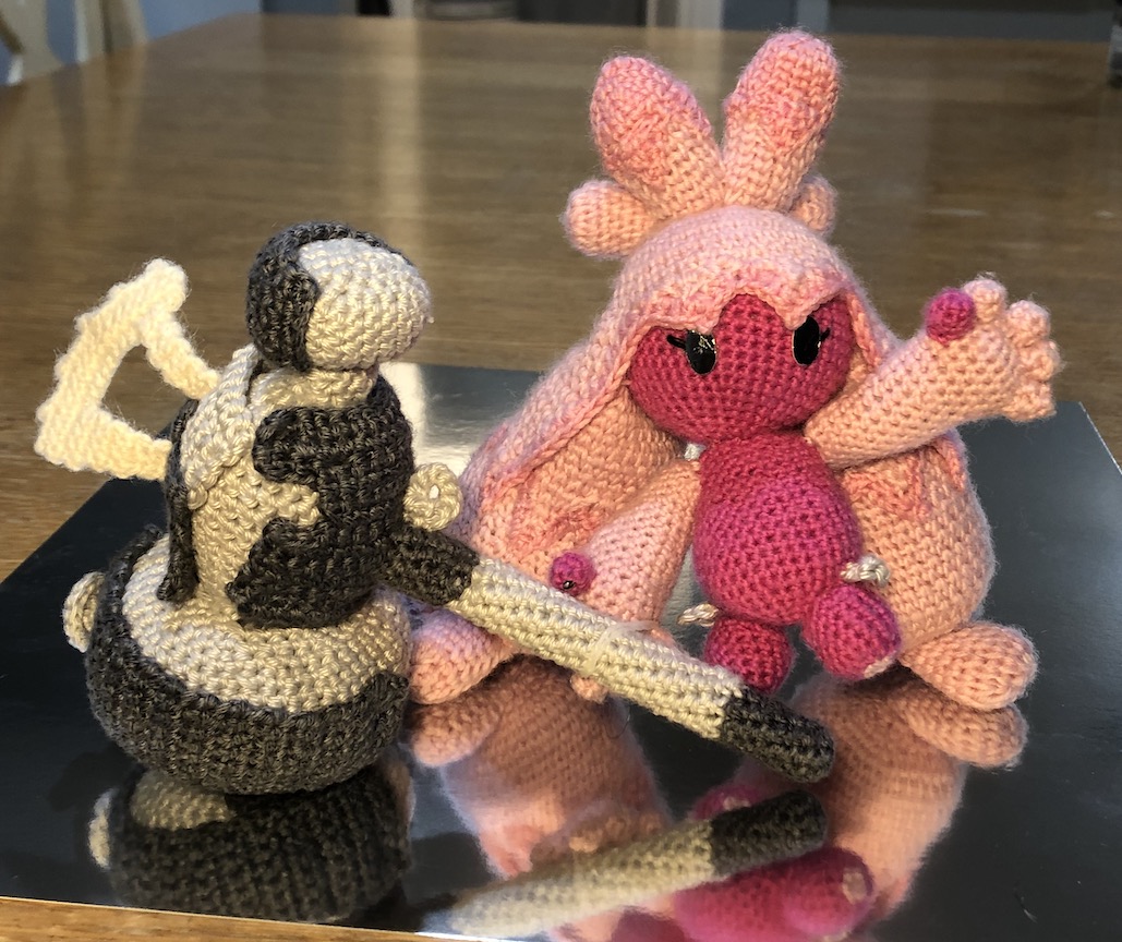 Crocheted Tinkaton standing on a reflective surface, one hand on her hammer, one in the air