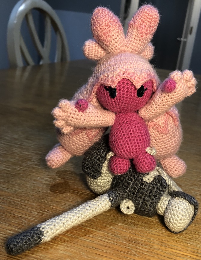 Crocheted tinkaton standing on top of her hammer laying on the ground. She has both hands out to keep balance