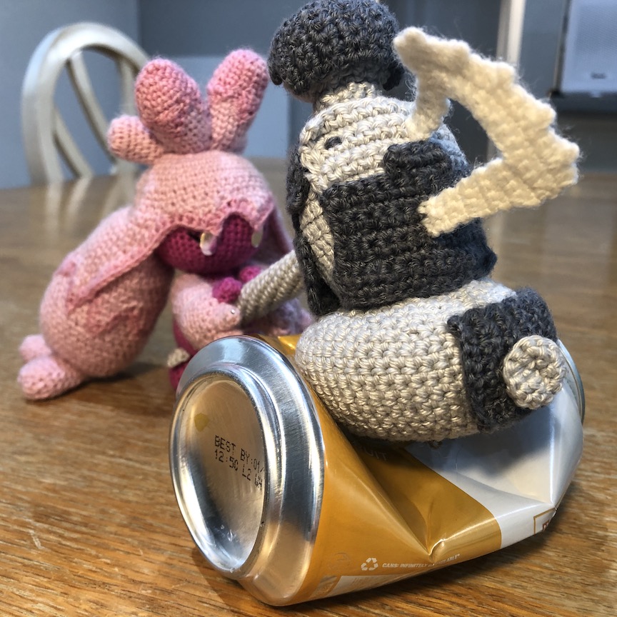 Crocheted Tinkaton with both hands on her hammer as she uses it to crush a soda can