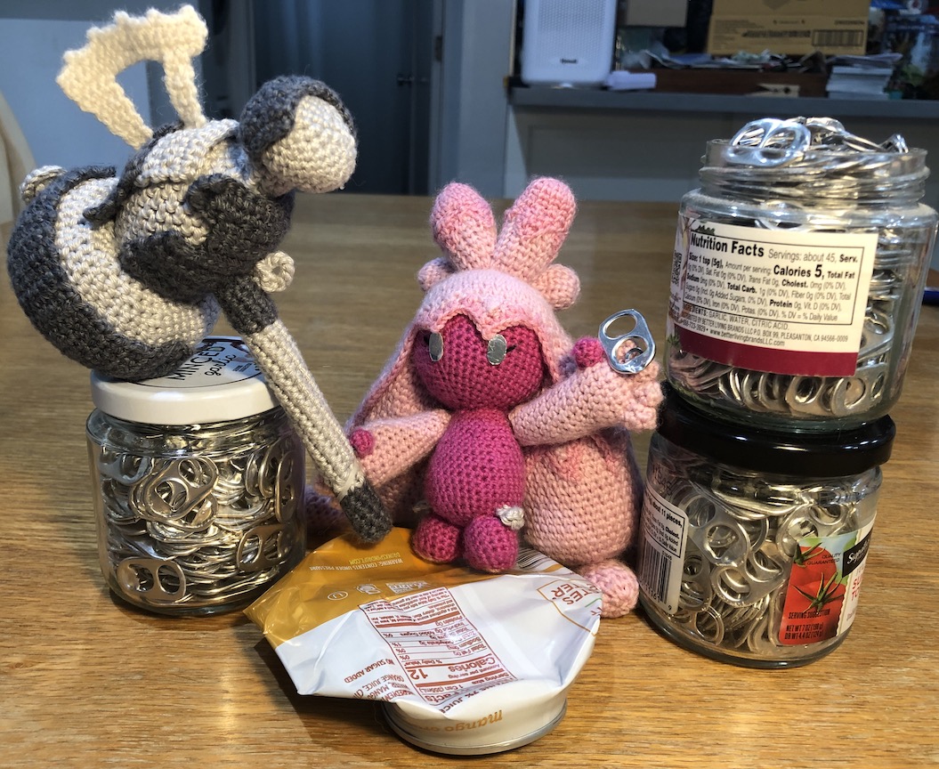 Crocheted Tinkaton proudly holding the pull tab from the soda can in one hand and her hammer in the other. She stands on top of the completely flattened can, and next to three jars FULL of pull tabs