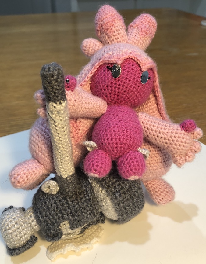 crocheted tinkaton standing on the base of her hammer, one hand holding onto the handle, sticking up perpendicular to the ground. She leaning to the side a little and her other arm is out to steady herself