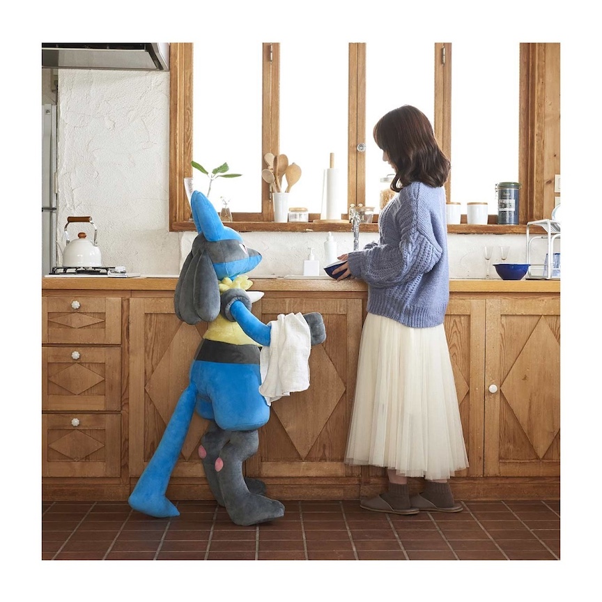Big Lucario plushie standing with a dishcloth next to a woman doing dishes