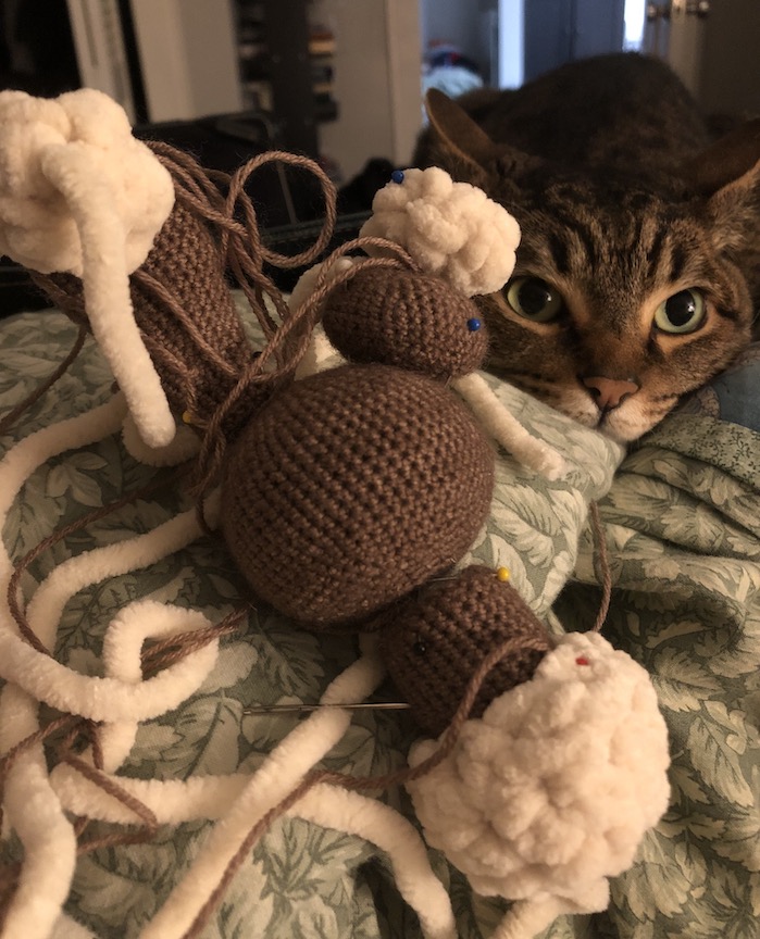 Mostly complete crocheted buneary, with no face or feet yet and the feet and ears pinned on. Charlie, the tabby cat, is resting his head next to the incomplete pokémon