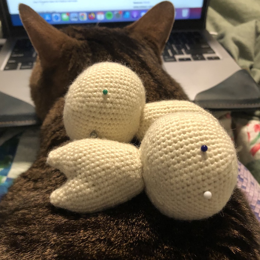 Two off-white crocheted little guys with orbs for heads, bodies with little cone shaped legs. They have pins for eyes and are laying on the back of a tabby cat