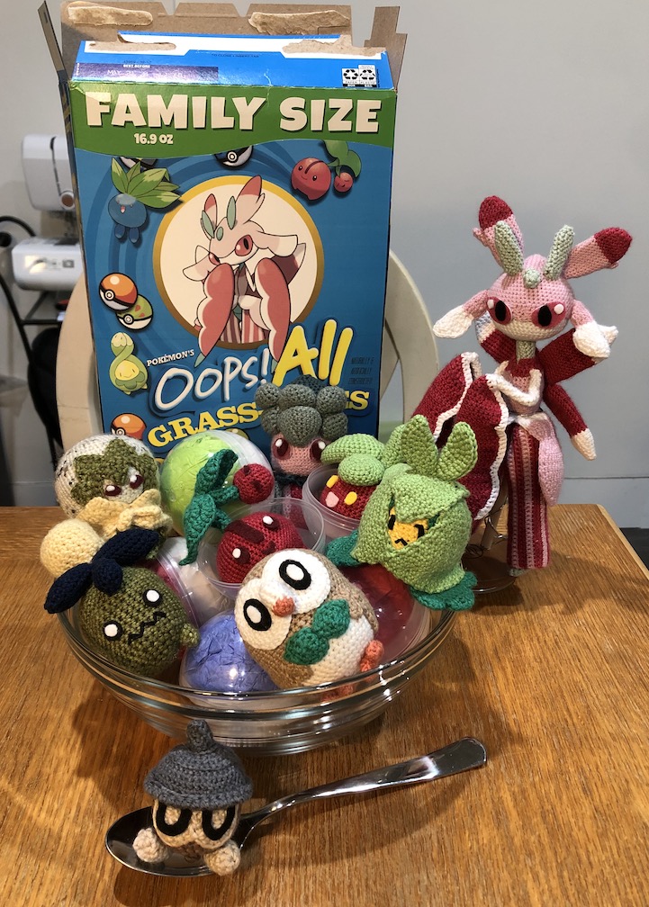 A custom Oops! All Berries cereal box has art with Grass-type pokémon and reads 'Oops! All Grass-Types.' Lurantis stands next to the box, and there's a glass bowl in front of it full of plastic pokéballs and the following crocheted pokémon: Smoliv, rowlet, eldegoss, cherubi, bounsweet, and swadloon. A spoon sits in front of the bowl with a crocheted seedot sitting in it