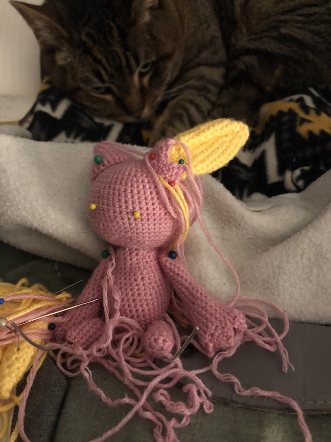 Partially finished shiny sneasel sits in front of an out-of-focus sleepy tabby cat. The unfinished sneasel has many strings from unattached pieces that are pinned to the body and the wire is sticking out through the footless legs. She has yellow pins standing in for her eyes