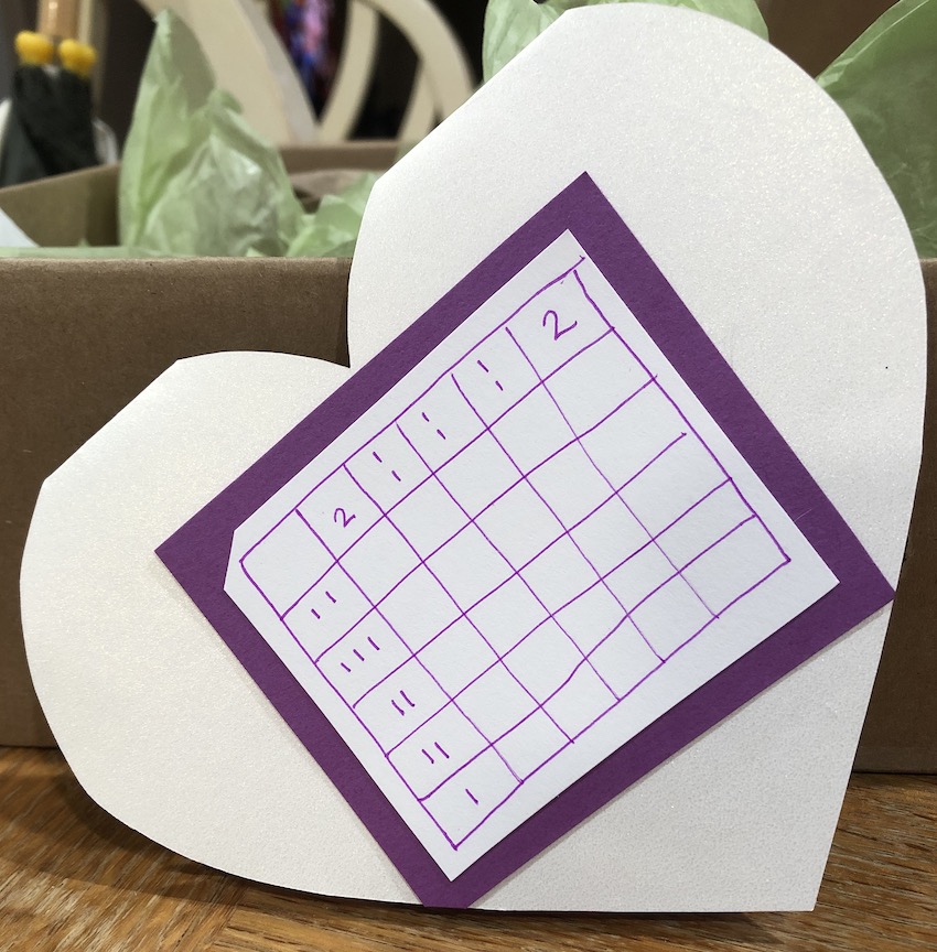 The back of the paper valentine with SCOUT on it, completely pearly white with a nonogram puzzle on it framed in a medium purple border