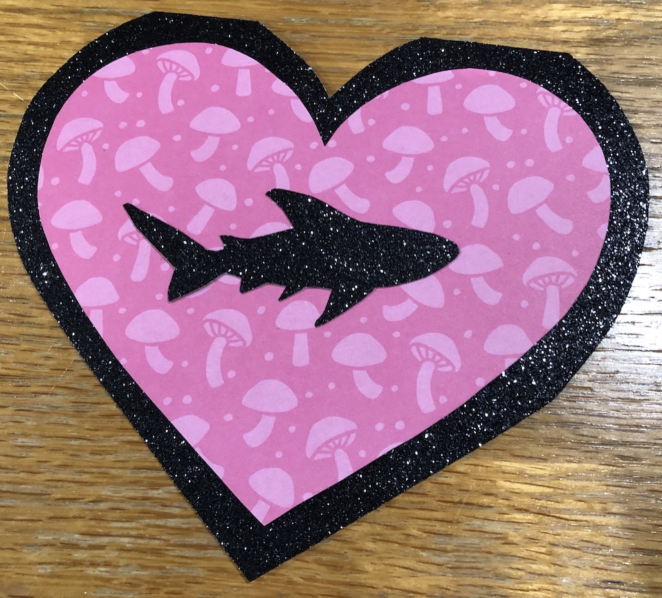 A heart shaped paper Valentine card with a black glitter border, hot pink mushroom patterened heart, and a shark cut out of the same black glitter cardstock