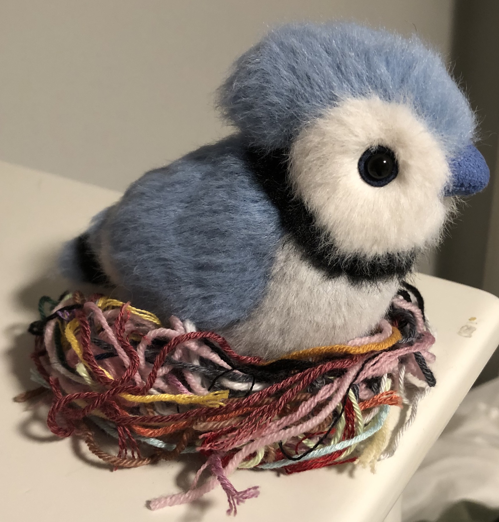 A little blue jay plushie sitting on a nest of assorted colorful yarn scraps
